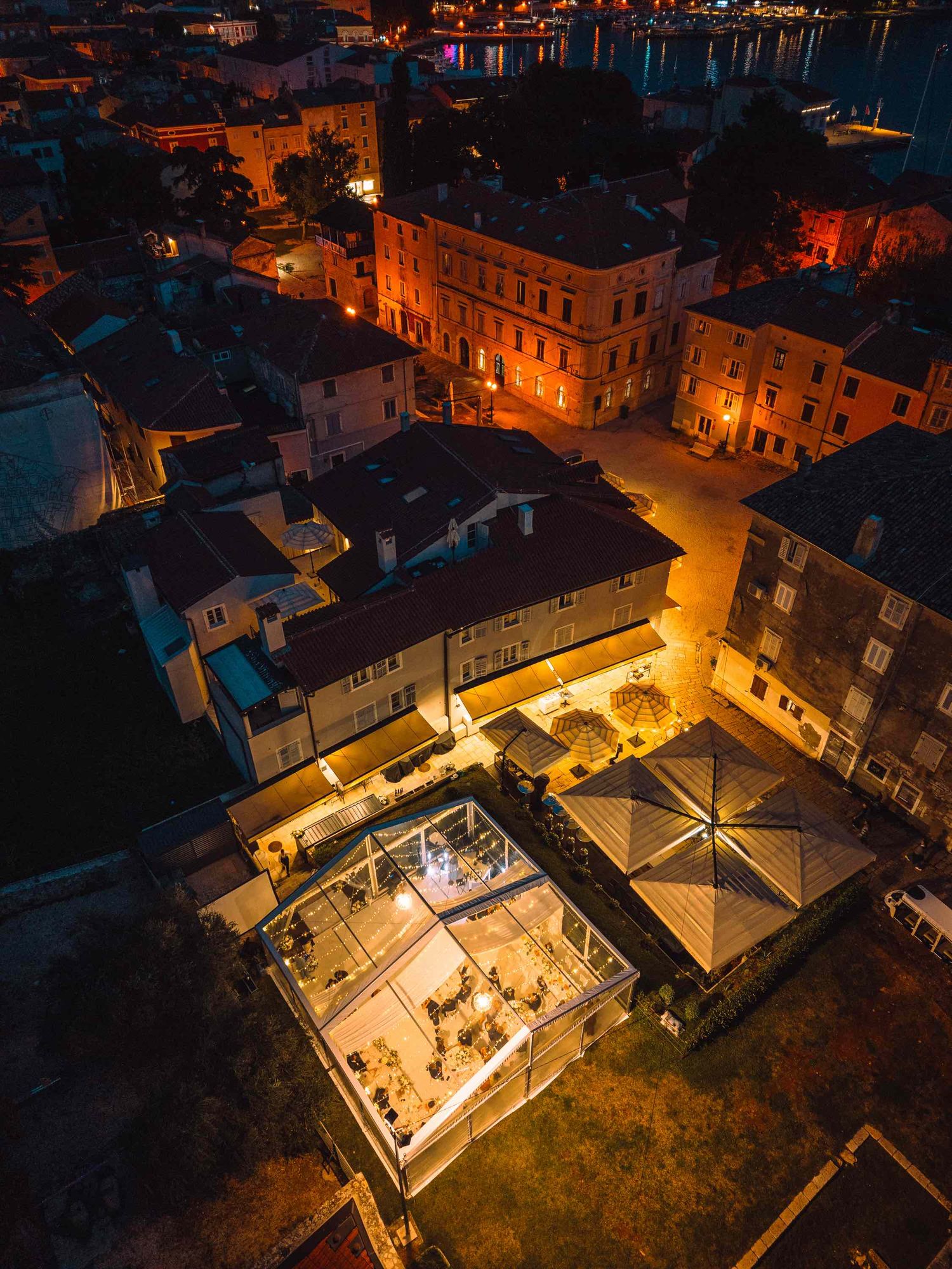 Aerial night view of a glowing glass restaurant courtyard surrounded by historic Mediterranean town buildings lit in warm amber light.