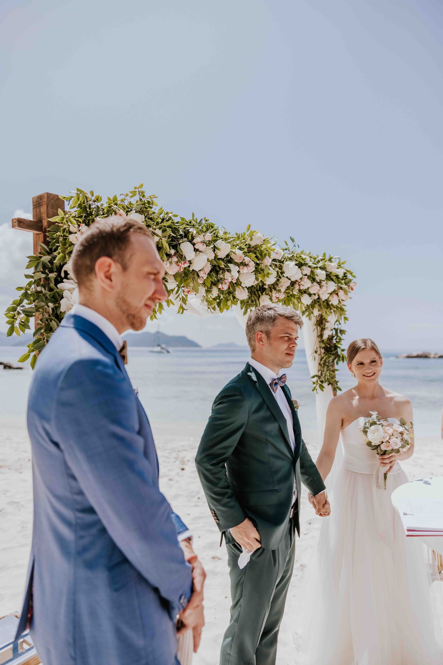 Bride and groom exchanging vows at beach wedding ceremony under floral arch.