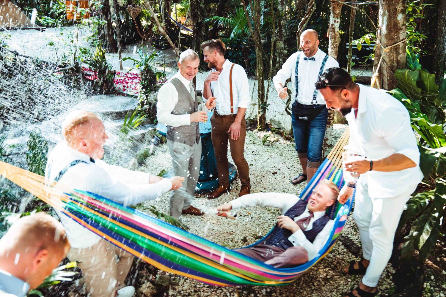 Groomsmen in suspenders relaxing in a colorful hammock outdoors in a tropical garden setting.