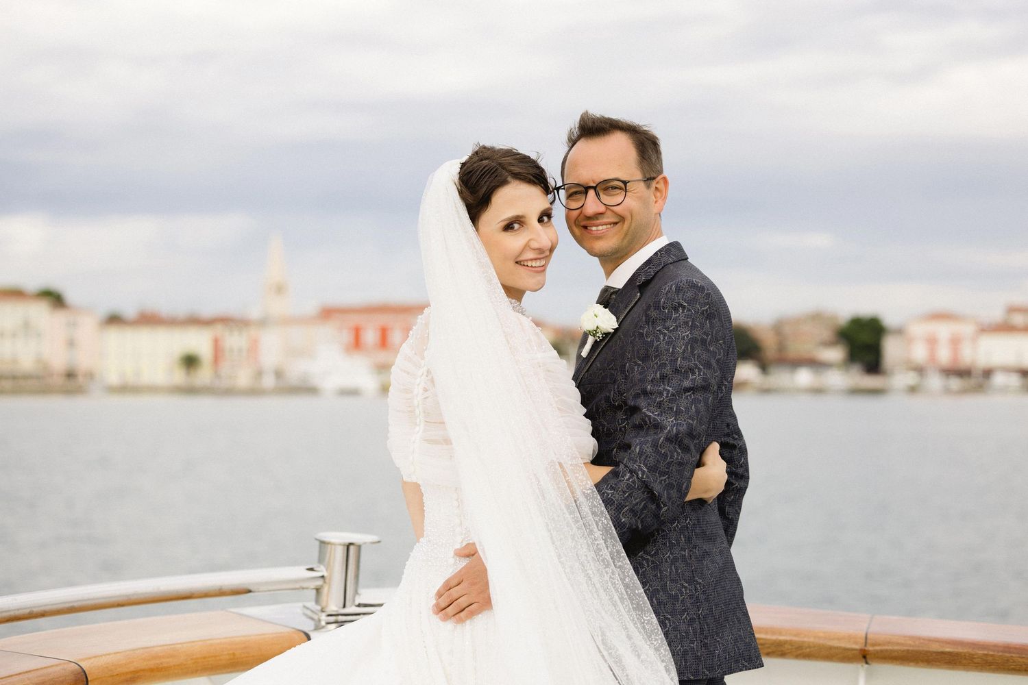 Smiling newlyweds embracing on a yacht with a picturesque harbor in the background.
