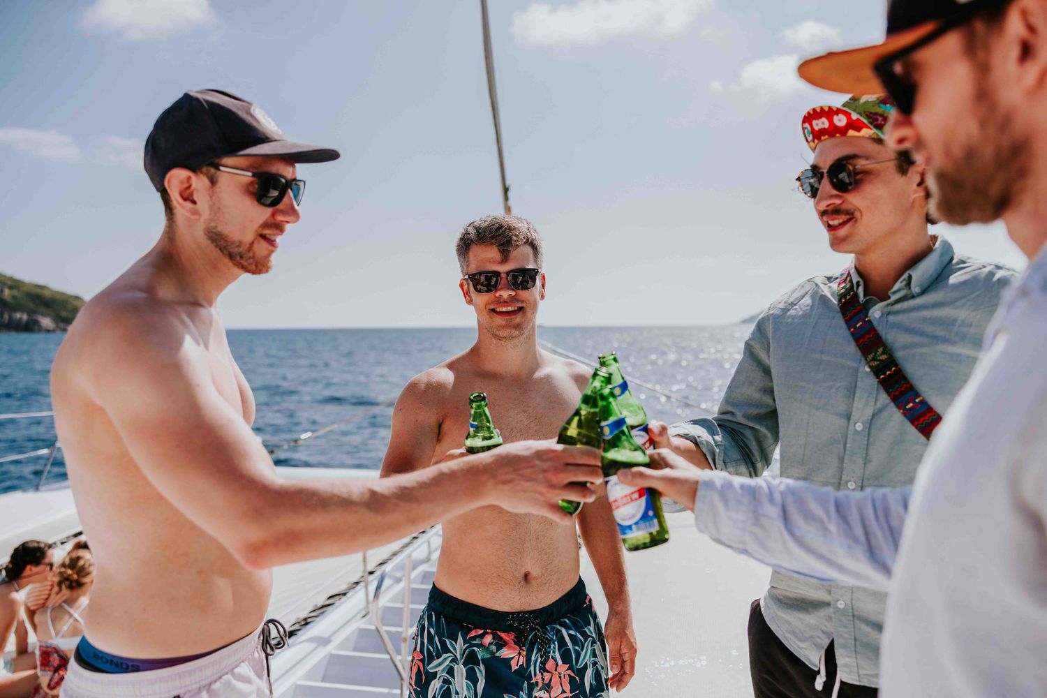Group of young men toasting with beer bottles on a sunny catamaran boat on the ocean.