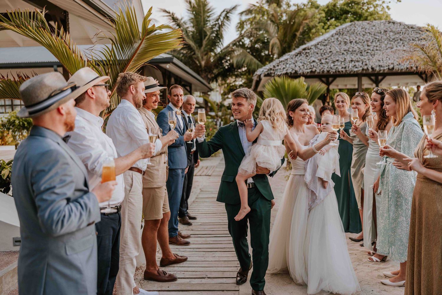 Groom carries bride through cheering wedding guests on a sunny tropical outdoor venue walkway.