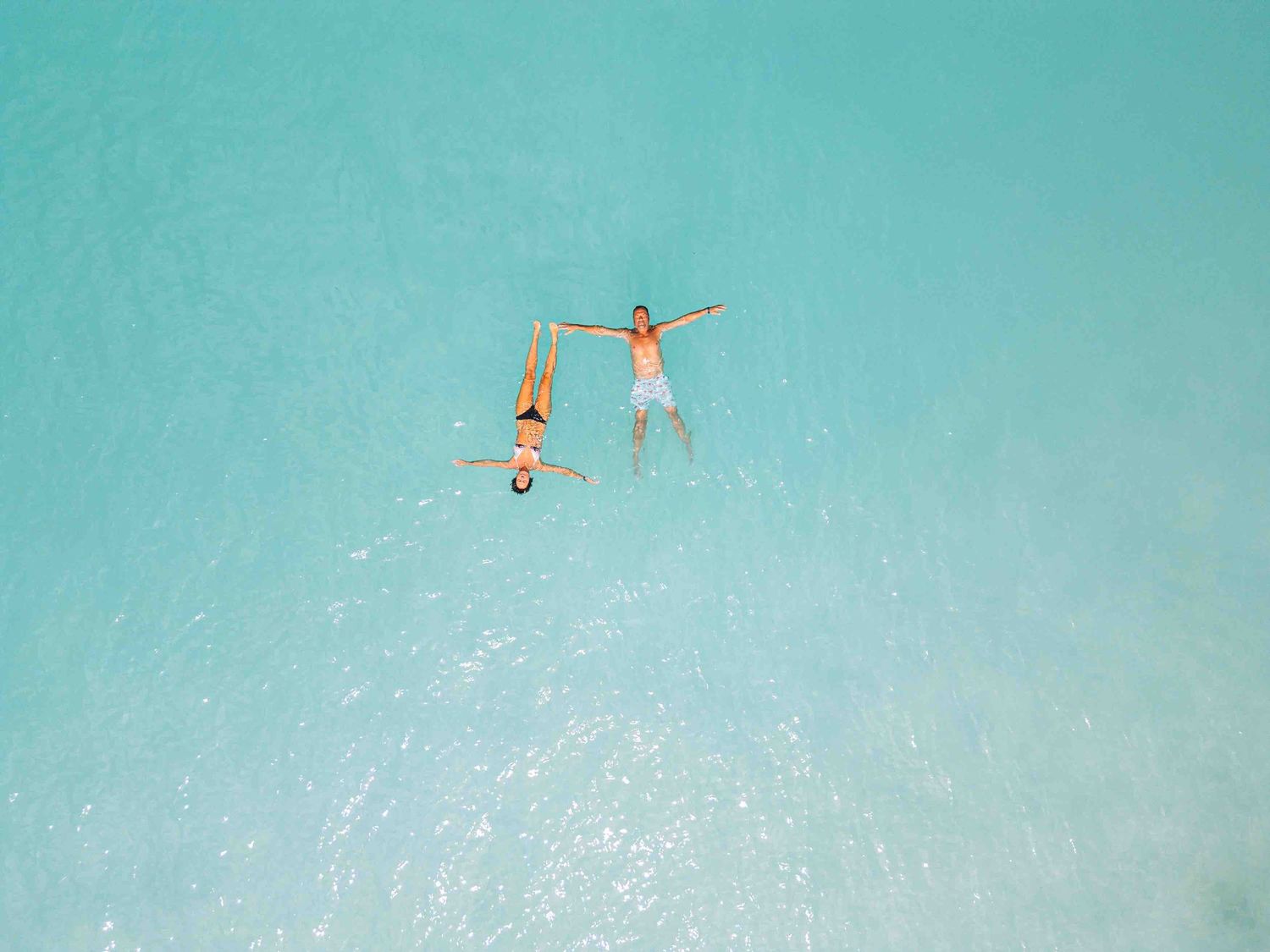 Aerial view of two people floating on their backs in clear turquoise tropical ocean water, holding hands.