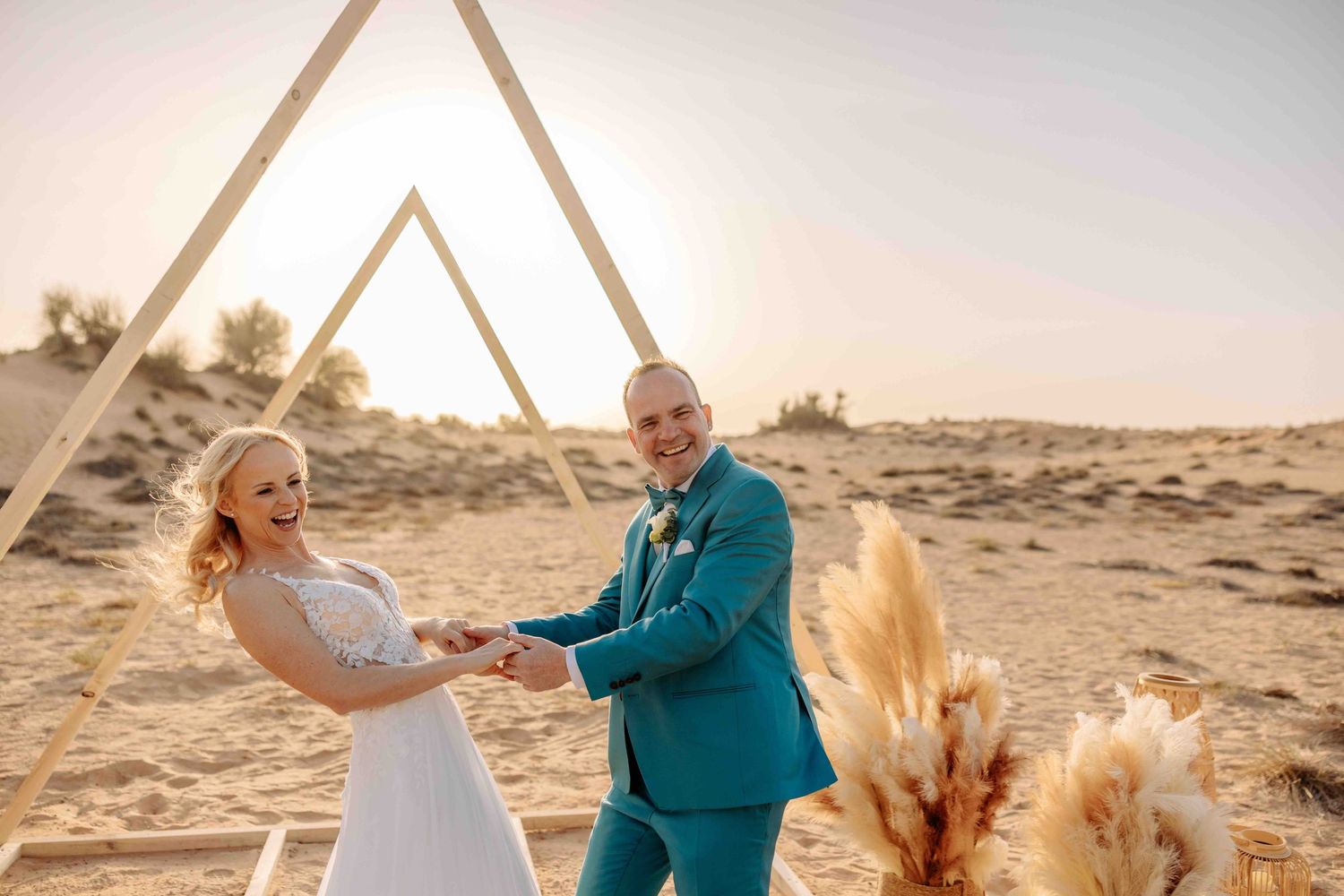 Happy couple exchanging rings at desert wedding with triangular pampas grass arch at golden sunset.