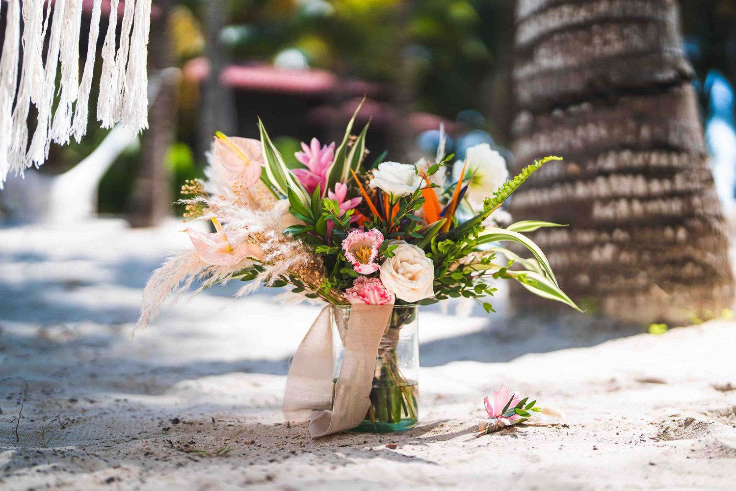 Tropical beach wedding bouquet with colorful flowers and ribbon on white sand near palm trees.