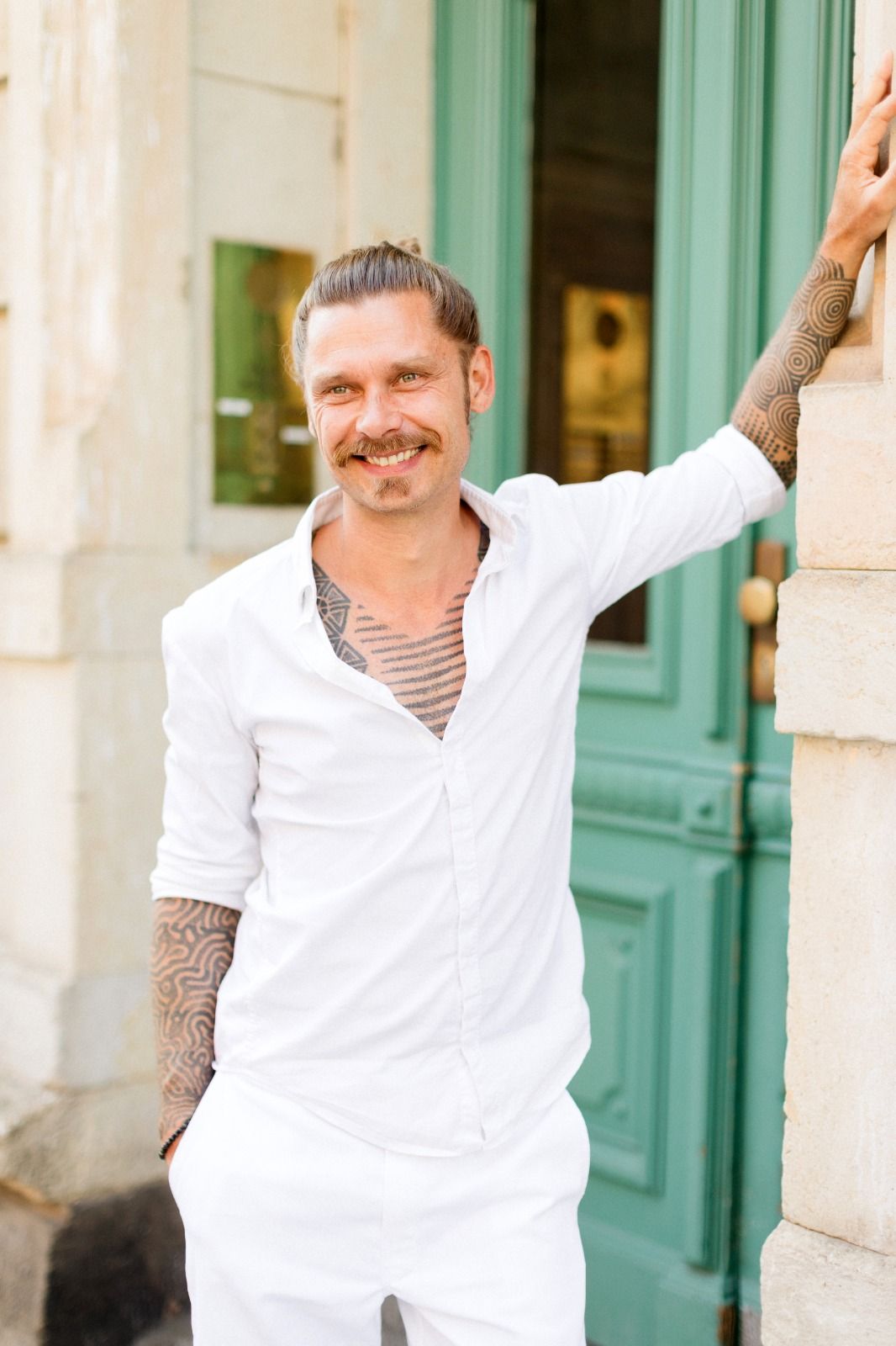 Smiling tattooed man in white shirt leaning against a green door in an urban setting.