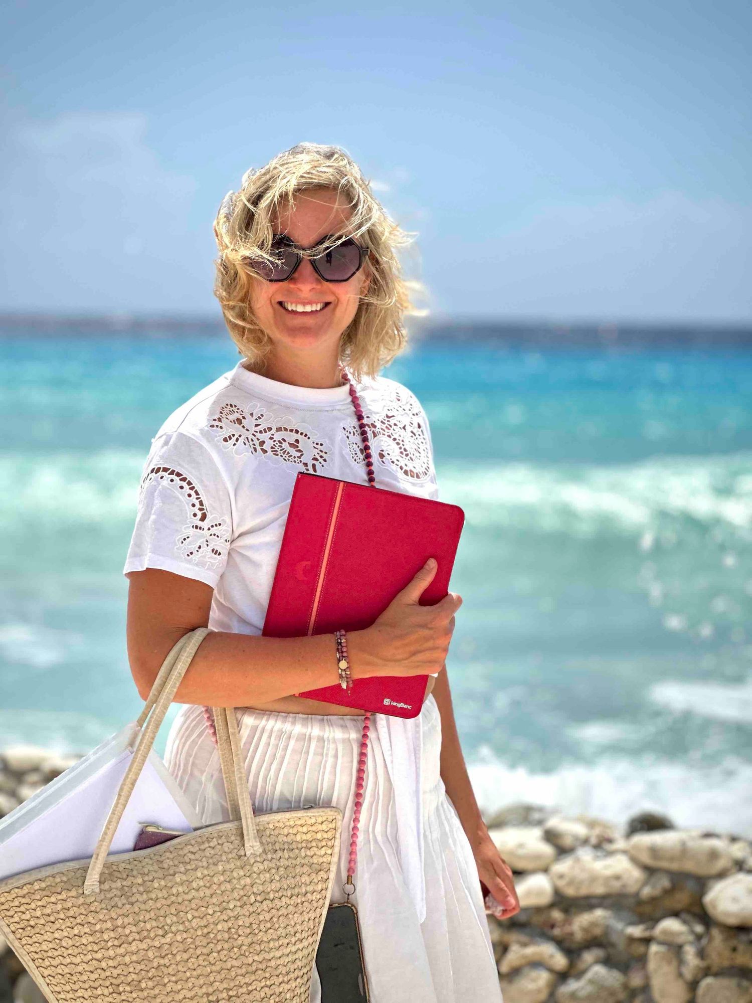 Smiling blonde woman in white dress holding a red book by the turquoise ocean on a sunny day.