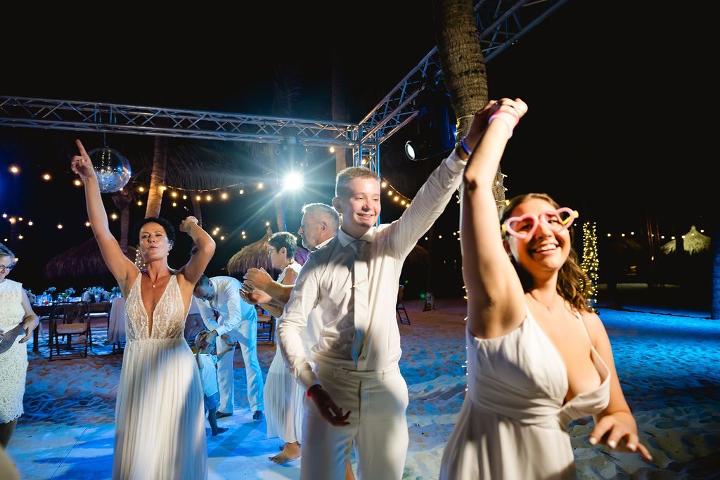 Guests dancing joyfully at an outdoor nighttime beach wedding reception under string lights.