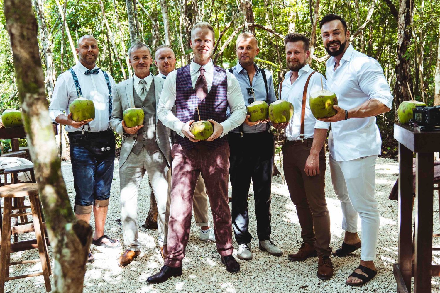 Groom and groomsmen in colorful suits holding coconut drinks in a tropical forest wedding venue.