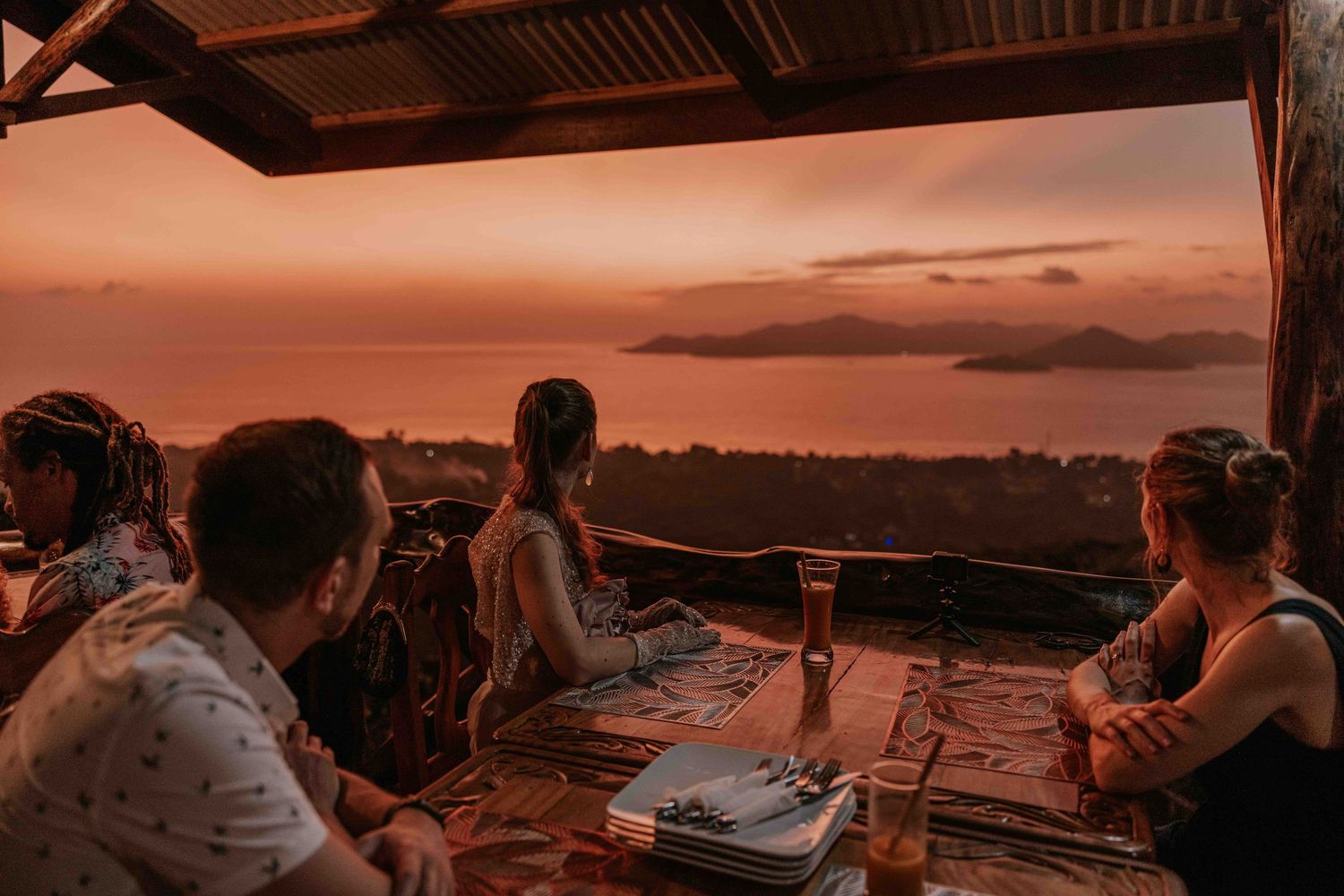 Friends dining on an open-air terrace with a stunning ocean sunset view in a tropical location.