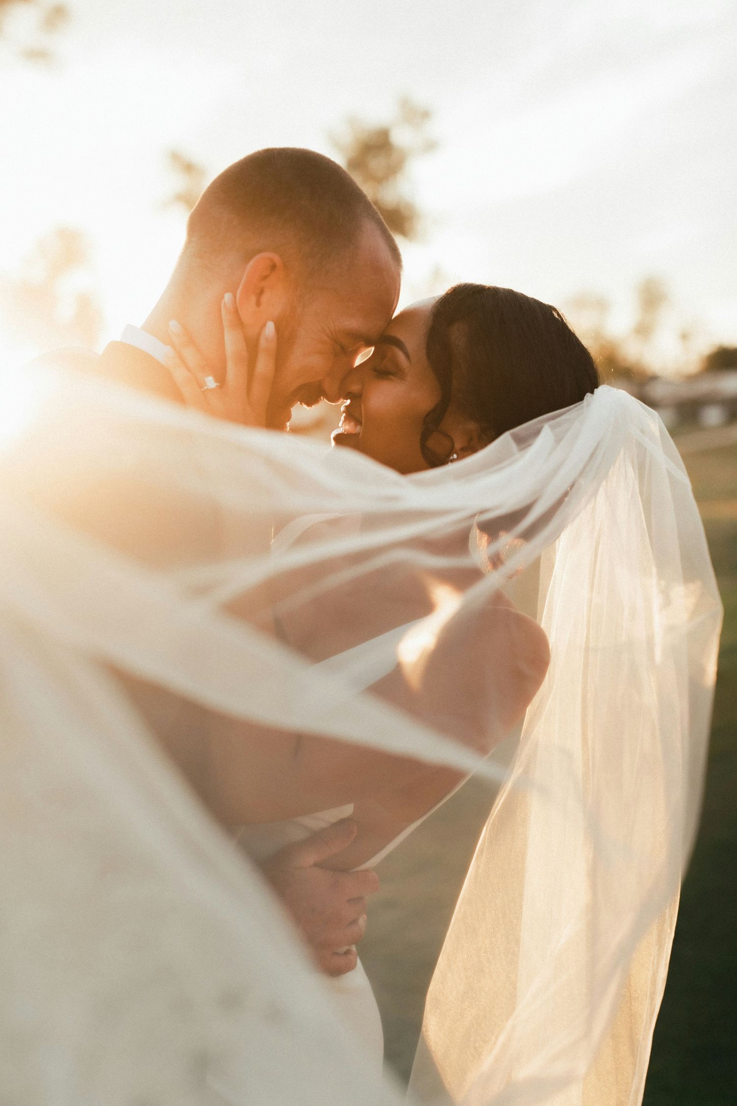 Romantic wedding couple sharing an intimate moment under a flowing veil in golden sunset light.