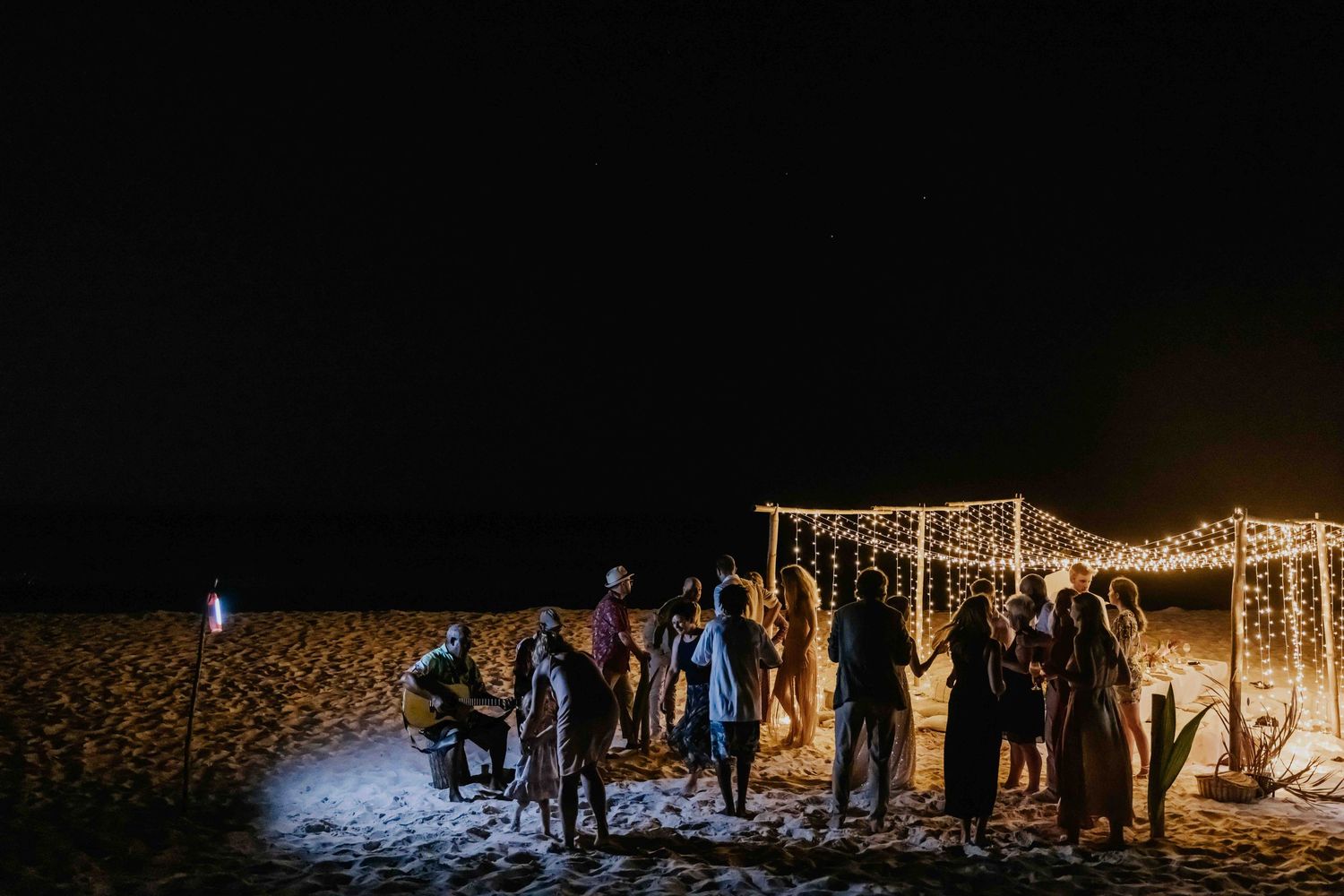Guests dancing and celebrating outdoors on beach at night near fairy light canopy.