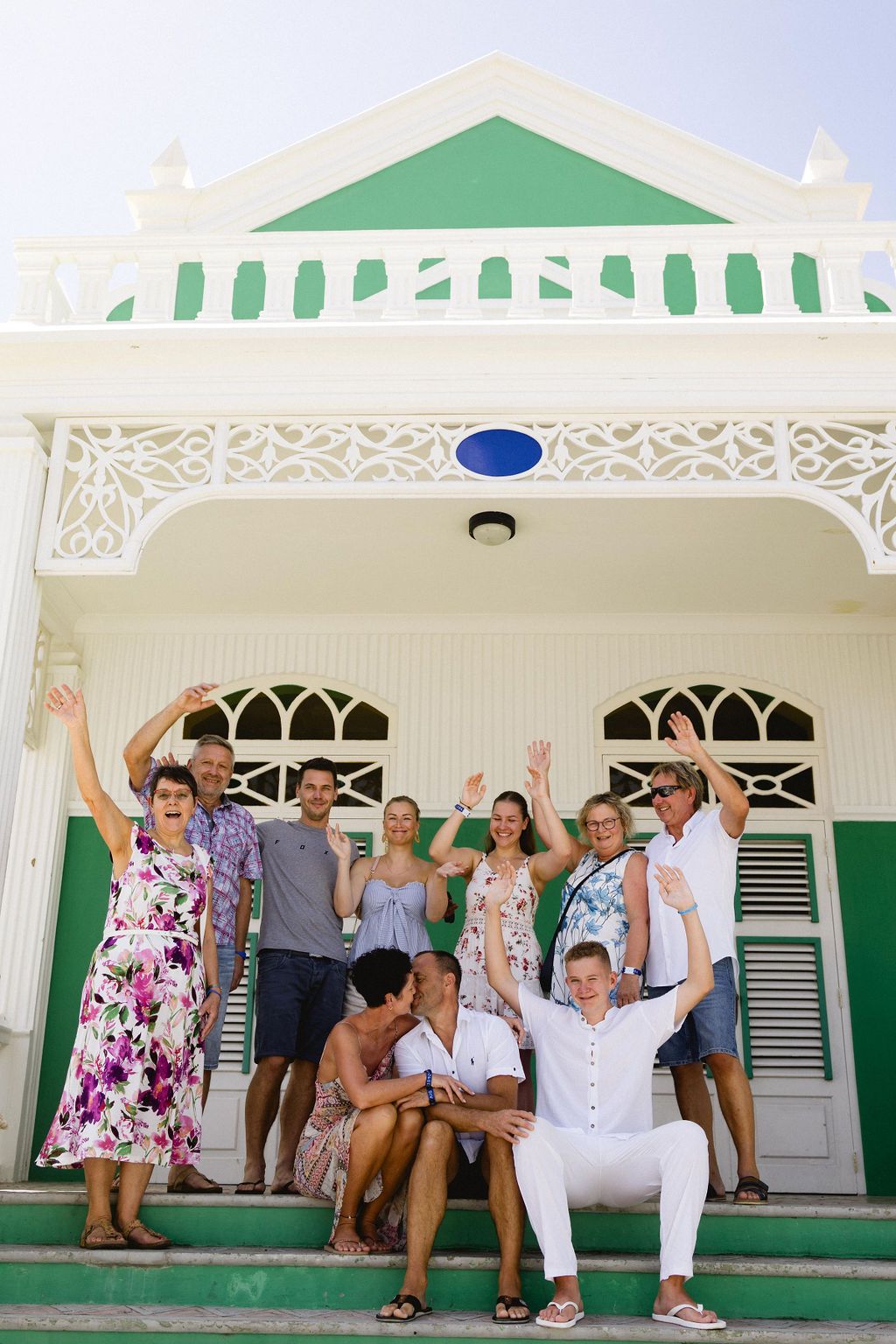 Cheerful group of wedding guests celebrate with raised arms on steps of colorful colonial building.
