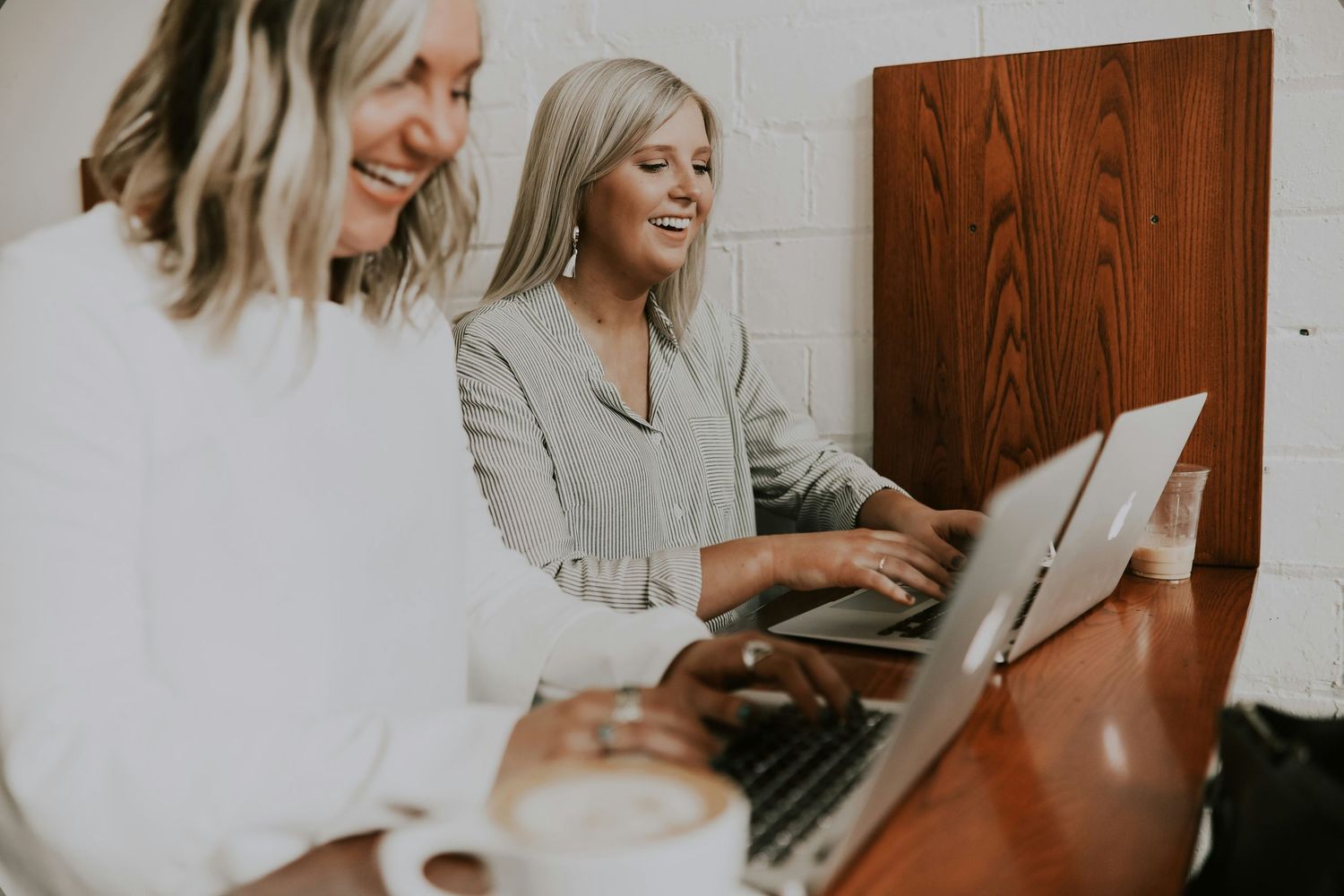 Two blonde women smile and work together on a laptop at a cafe table with coffee cups nearby.