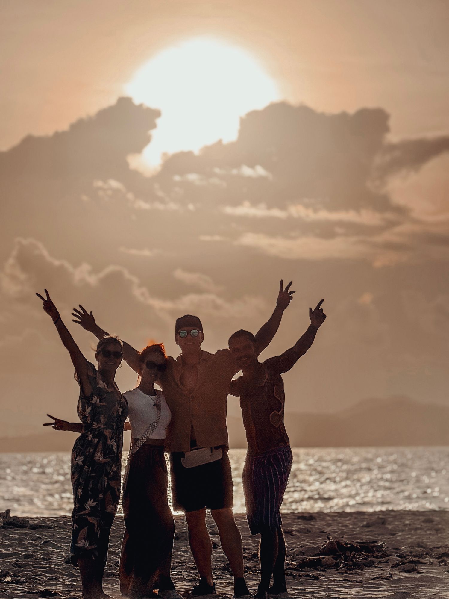 Group of four friends posing on a beach at sunset, silhouetted against a glowing golden sky.