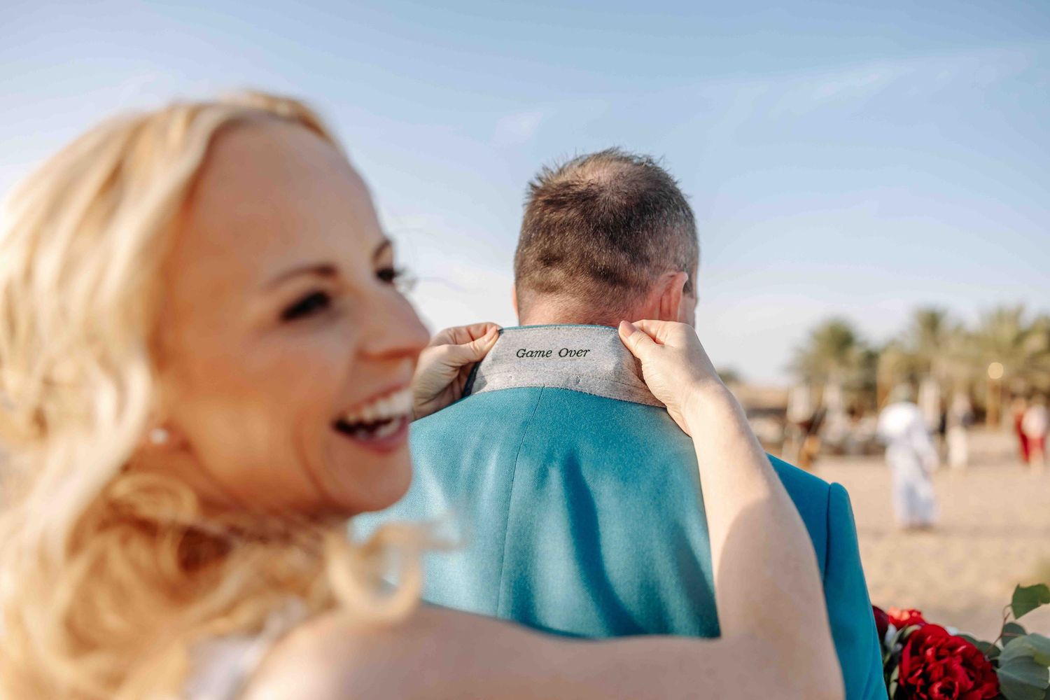 Smiling bride hugging groom in teal suit revealing hidden label message on collar at beach wedding.