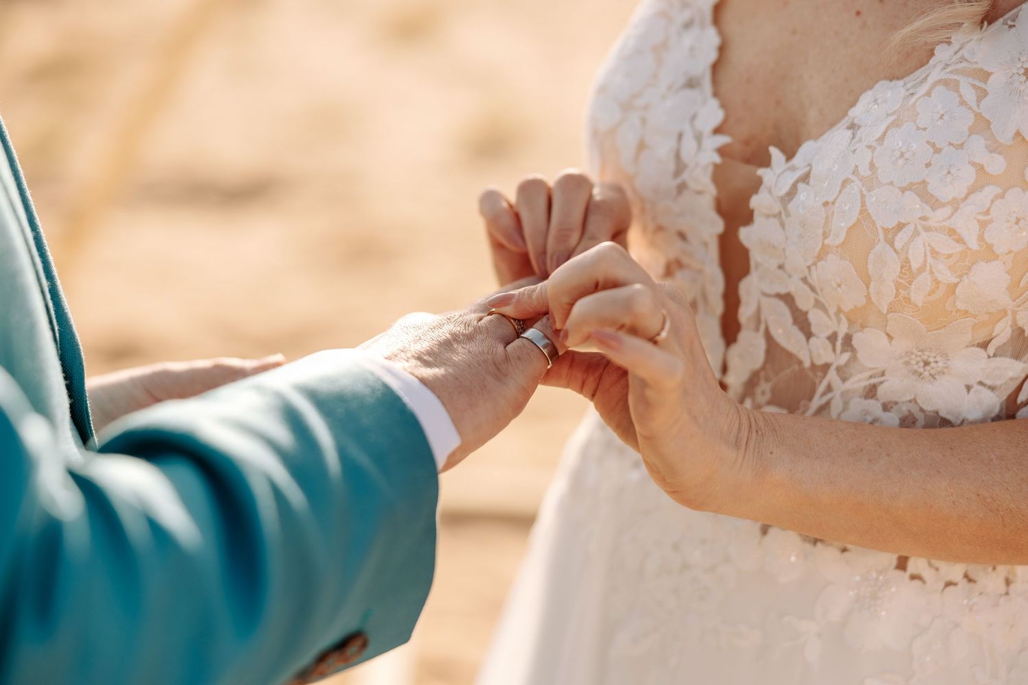 Groom in turquoise suit sliding wedding ring onto brides finger at sandy outdoor venue.