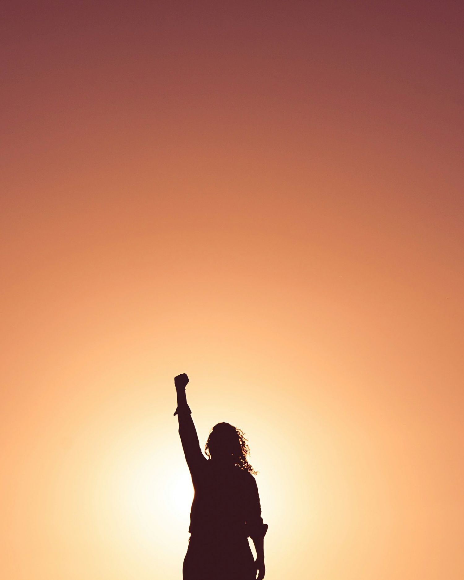 Silhouette of woman raising fist in triumph against warm golden sunset sky.