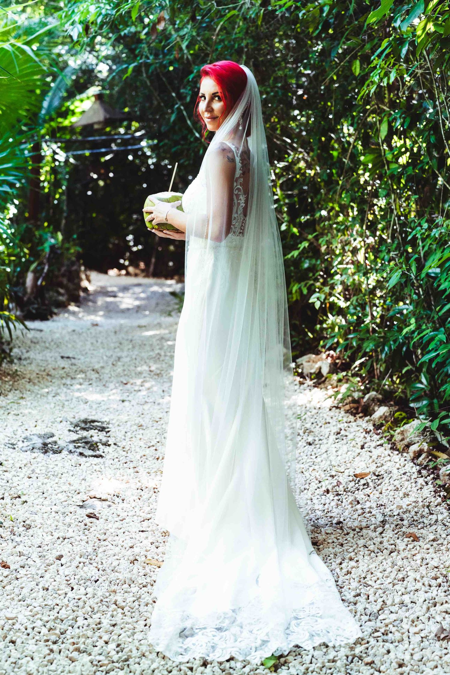 Bride in white lace gown and veil holding a coconut drink in a lush tropical garden setting.