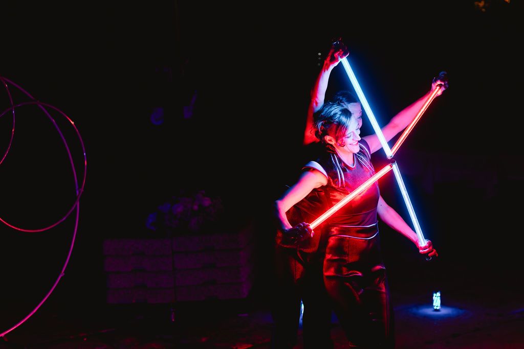 Female performer dancing with glowing neon light sabers in blue and red at dark nighttime event.