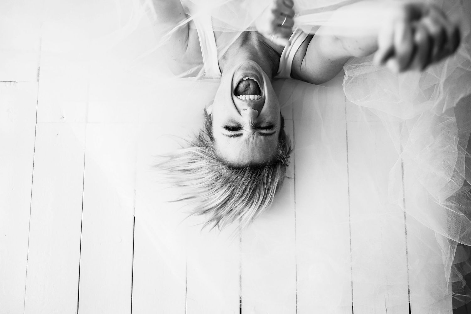 Black and white portrait of a laughing child lying upside down on white wooden floor.