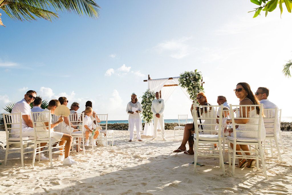 Intimate beach wedding ceremony with guests seated on white chairs under blue skies.