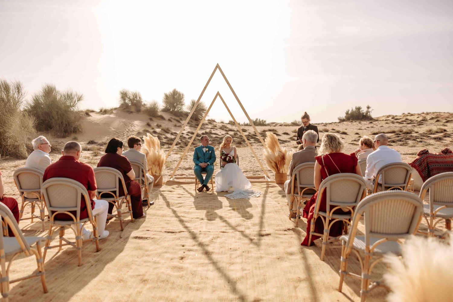 Intimate desert wedding ceremony with guests seated on rattan chairs facing triangular arch on sand dunes.