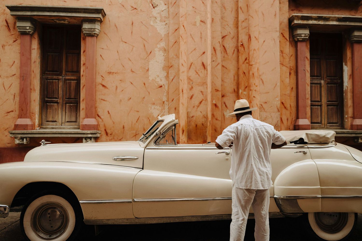 Man in white suit standing beside classic vintage cream convertible car on street.