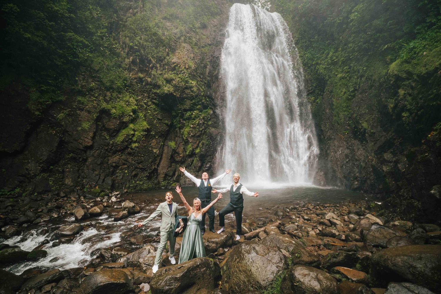 Wedding party celebrating joyfully in front of a majestic waterfall surrounded by lush green cliffs.