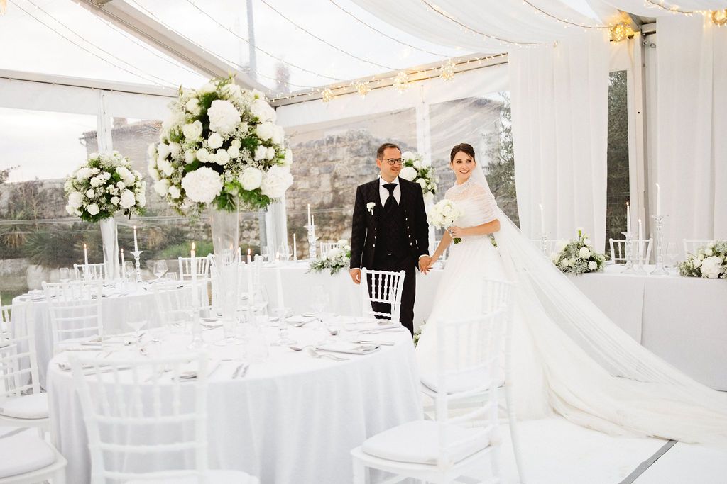 Bride and groom standing in an all-white wedding reception tent adorned with tall floral centerpieces.