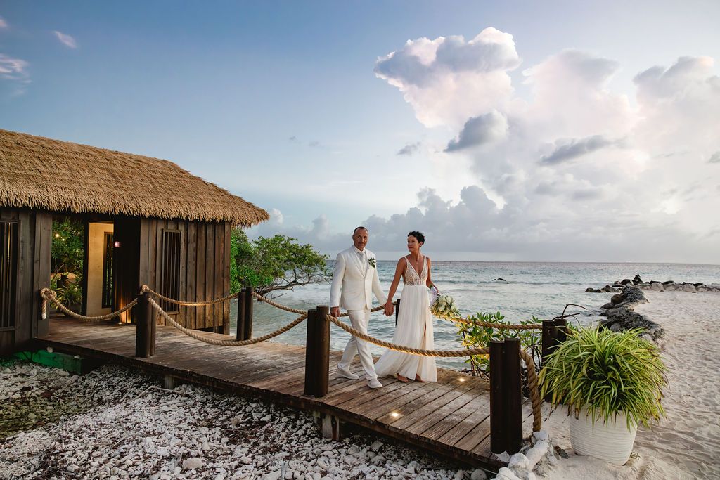 Newlywed couple walks hand-in-hand on wooden dock beside thatched bungalow on tropical beach resort.
