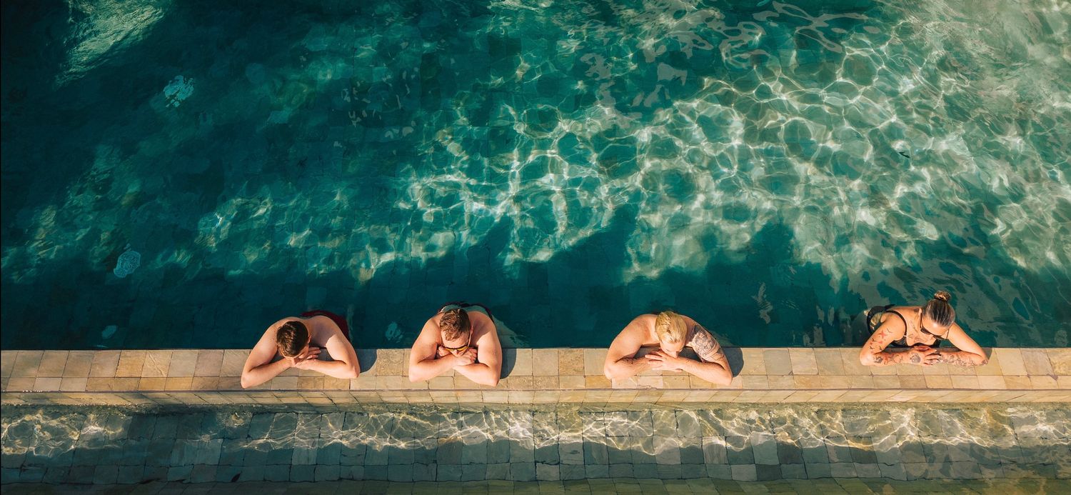 Four people relaxing at the edge of a turquoise swimming pool, leaning over the side on a sunny day.