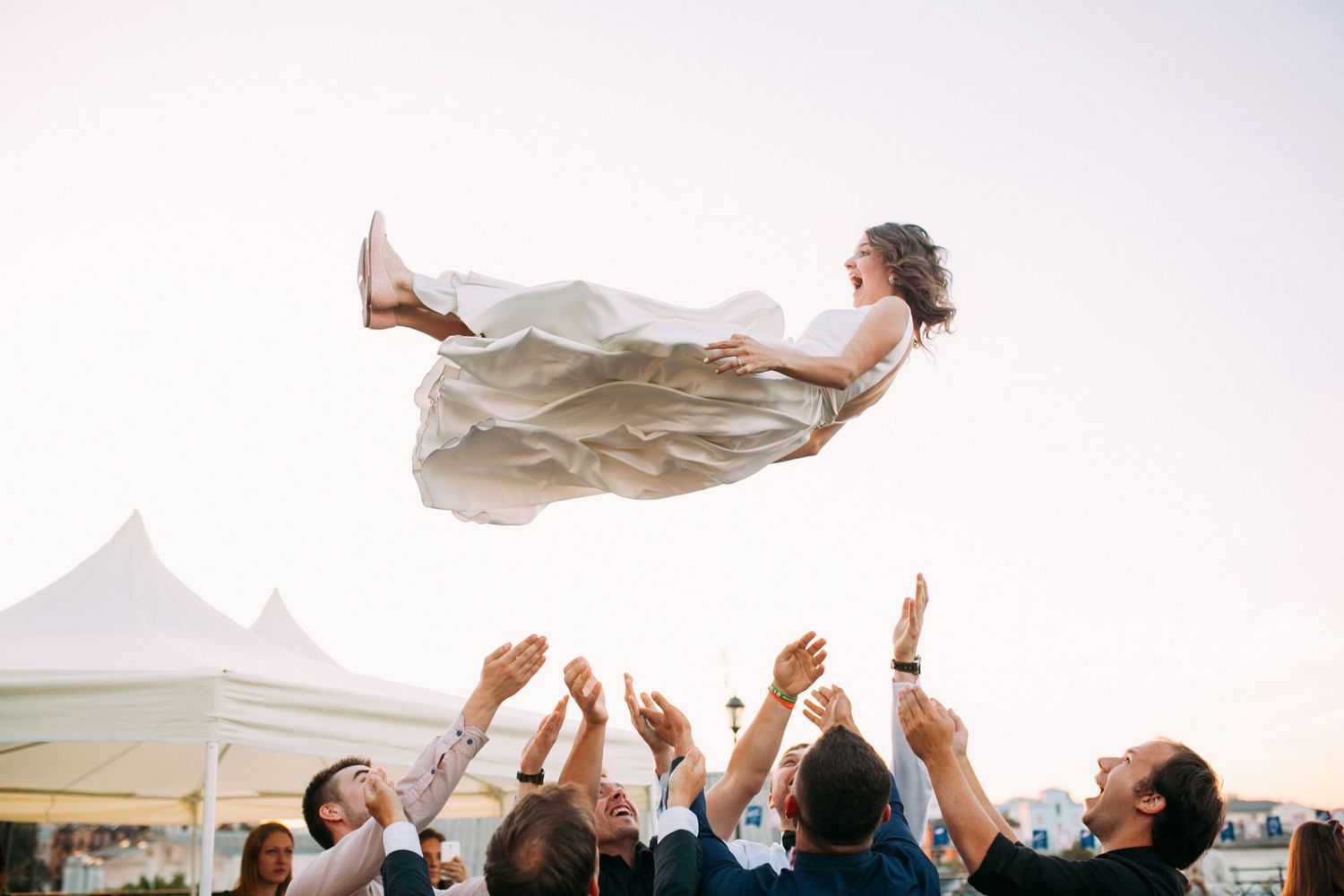 Bride being tossed in the air by wedding guests during outdoor reception celebration.