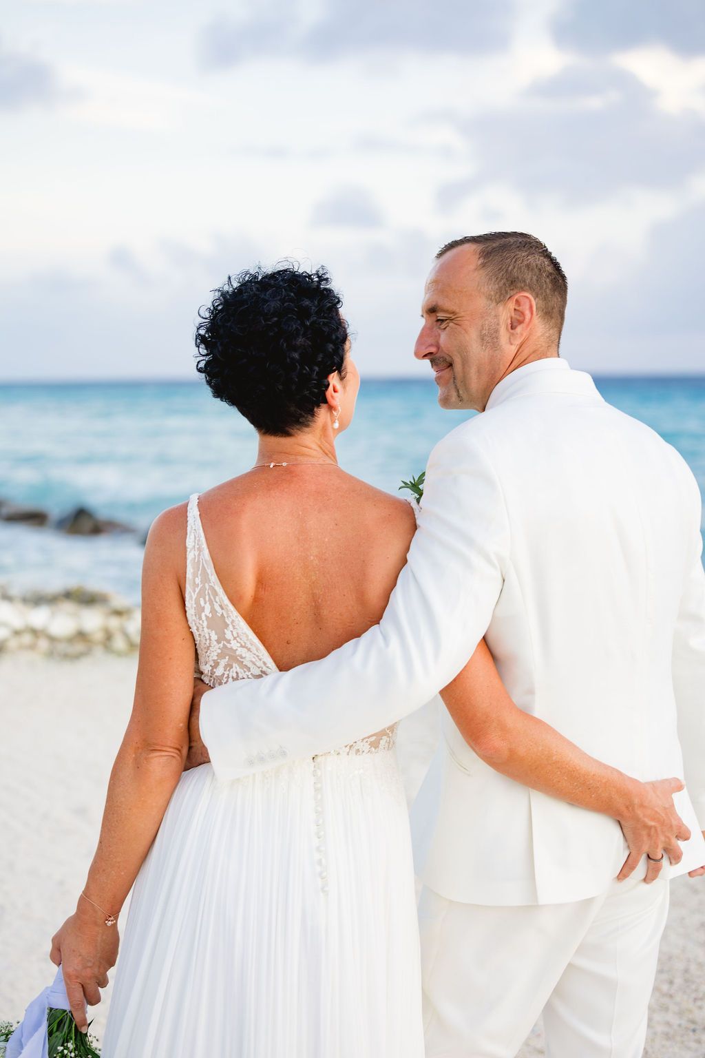 Bride and groom in white standing together on a beach facing the turquoise ocean.