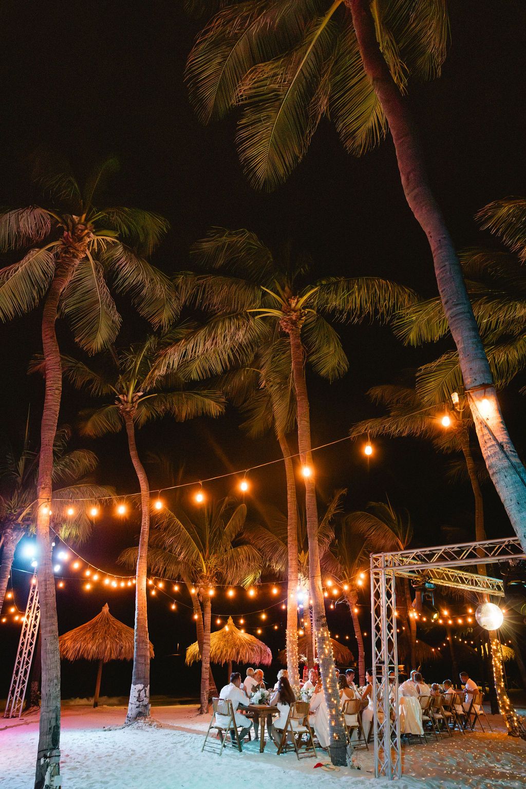 Romantic outdoor beach wedding reception dinner under string lights and tall palm trees at night.