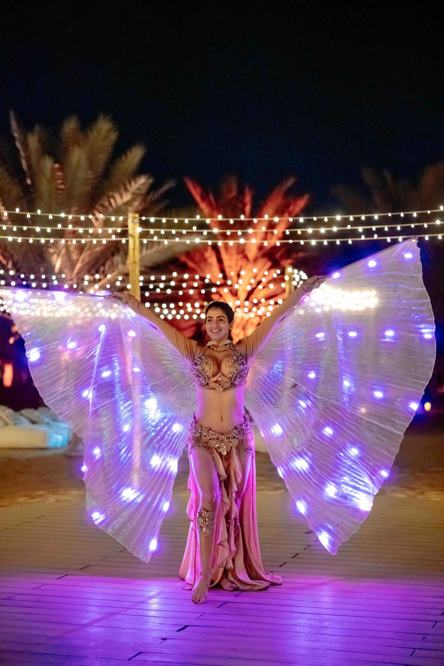 Belly dancer performing with illuminated LED wings costume at night under string lights and palm trees.