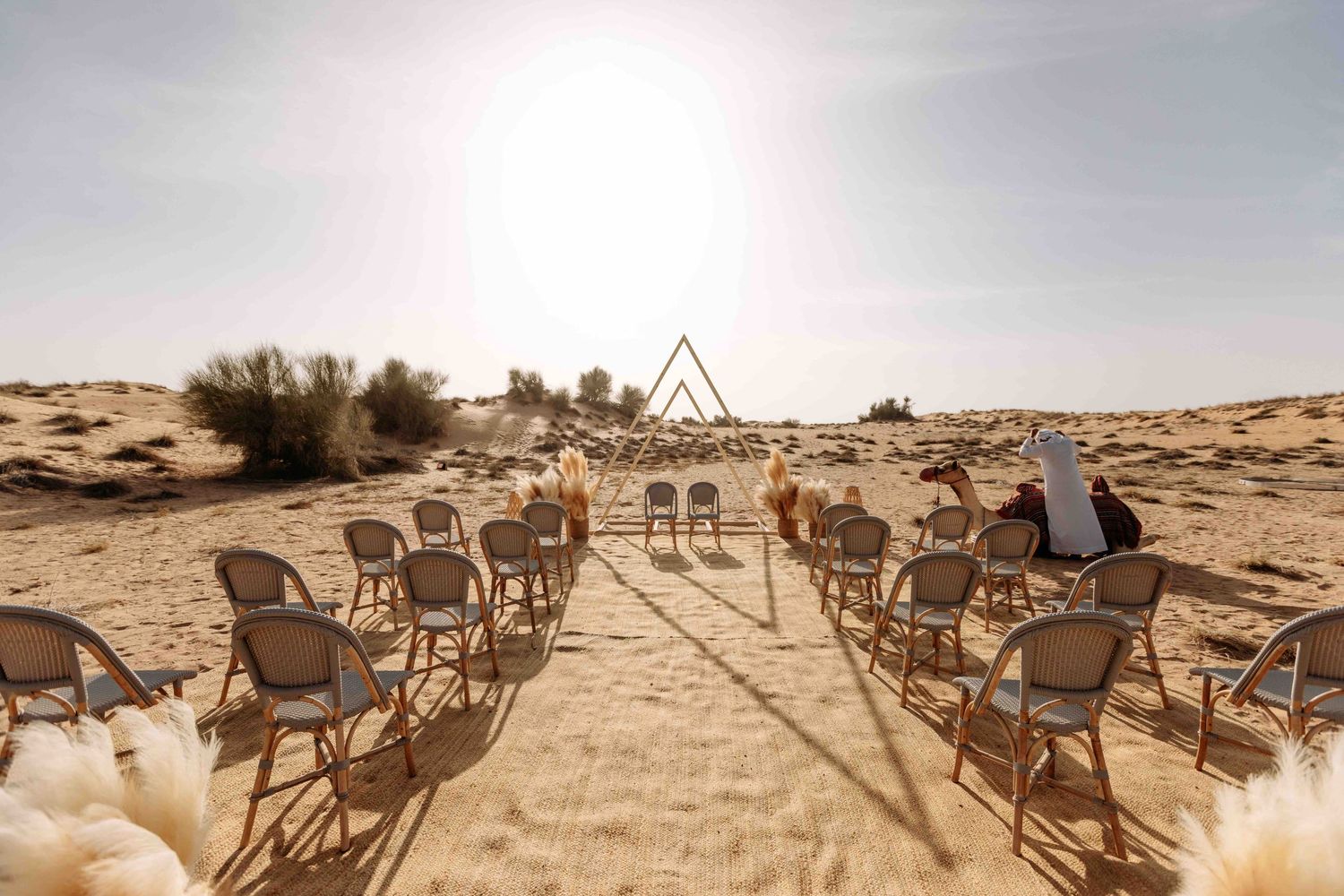 Outdoor desert wedding venue with rattan chairs and geometric arch in sandy landscape.