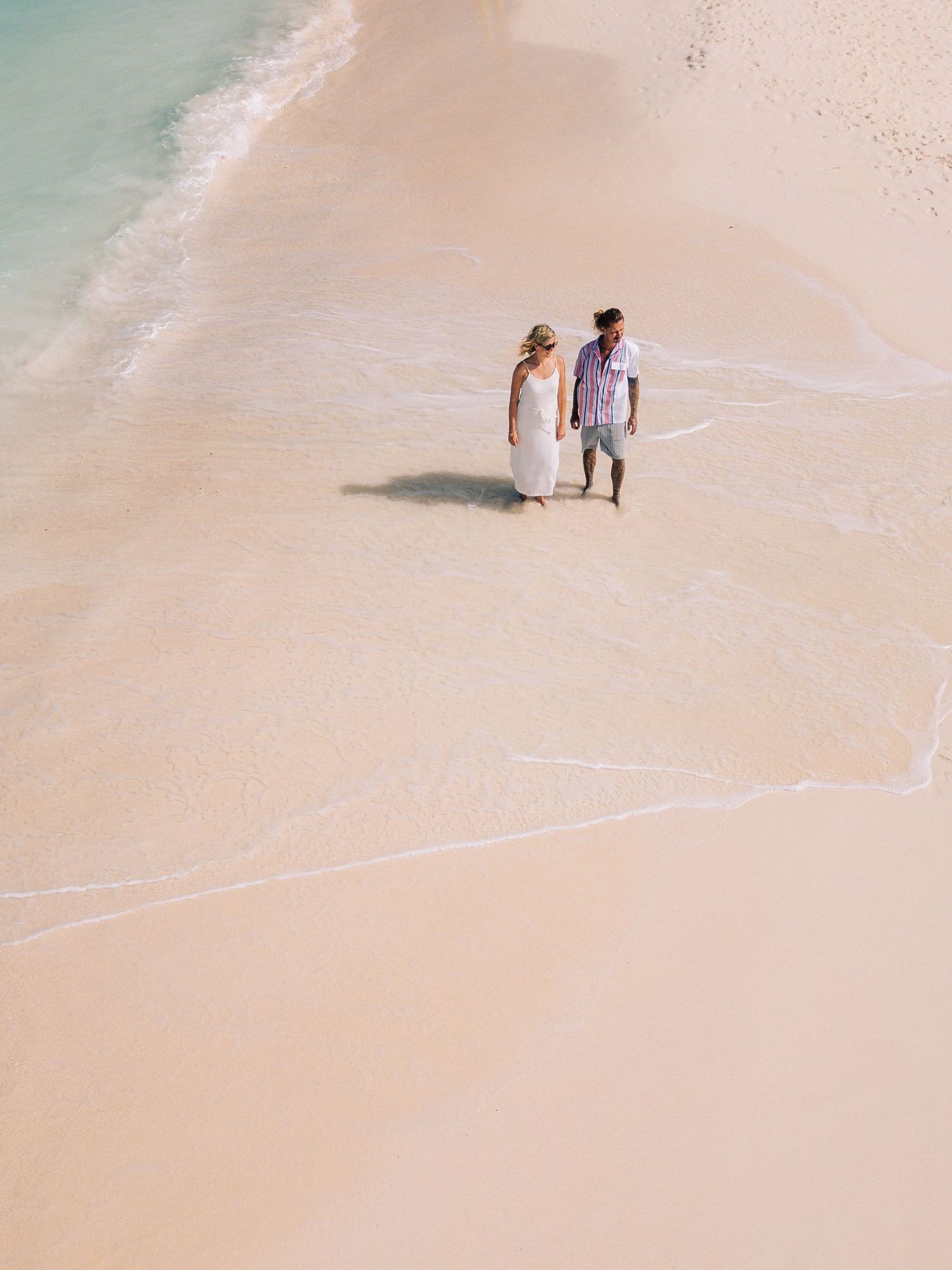Aerial view of a couple in wedding attire walking hand in hand on a pink sand beach with gentle waves.