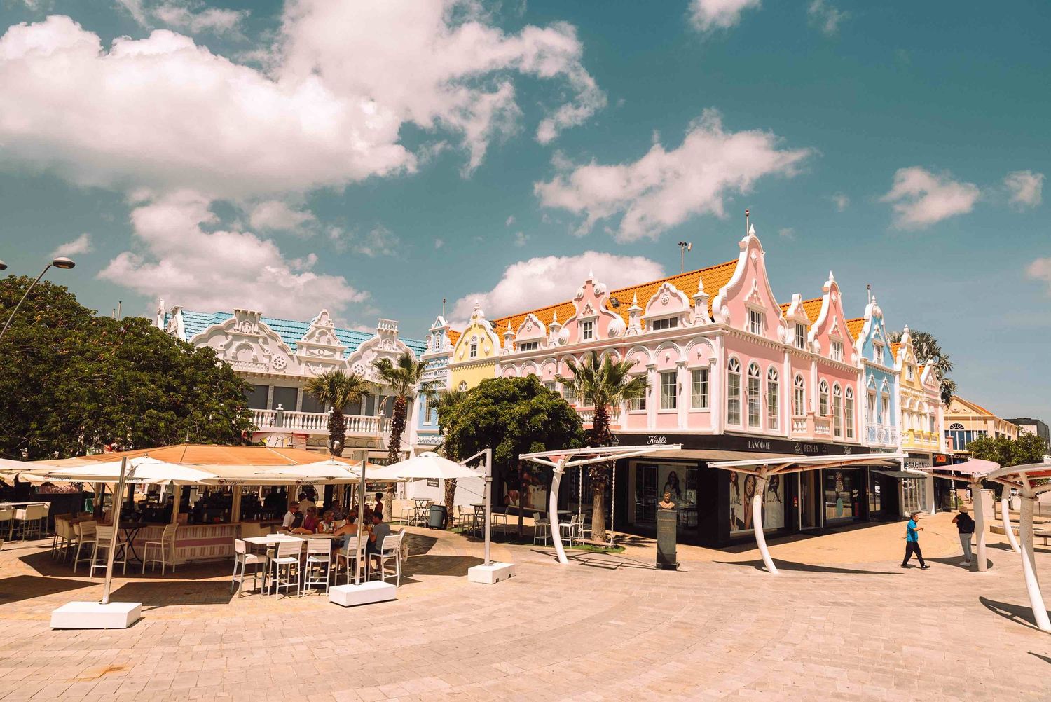 Colorful Dutch colonial architecture lines a sunny waterfront promenade in Oranjestad, Aruba, under a blue sky.