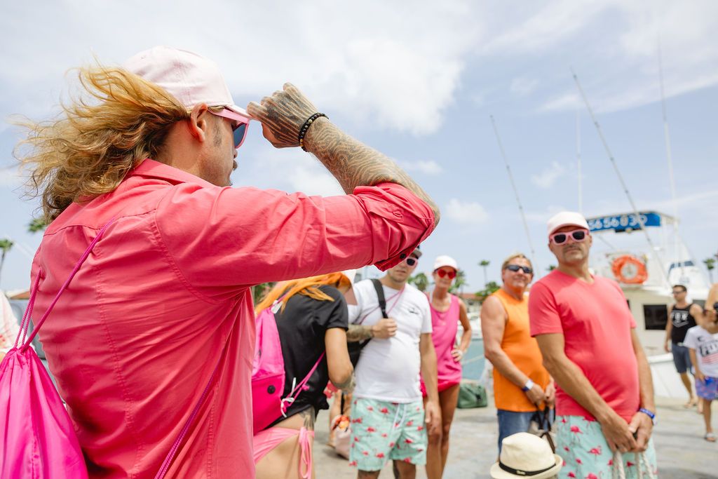 Woman in pink outfit shielding eyes from wind at a sunny marina with group.