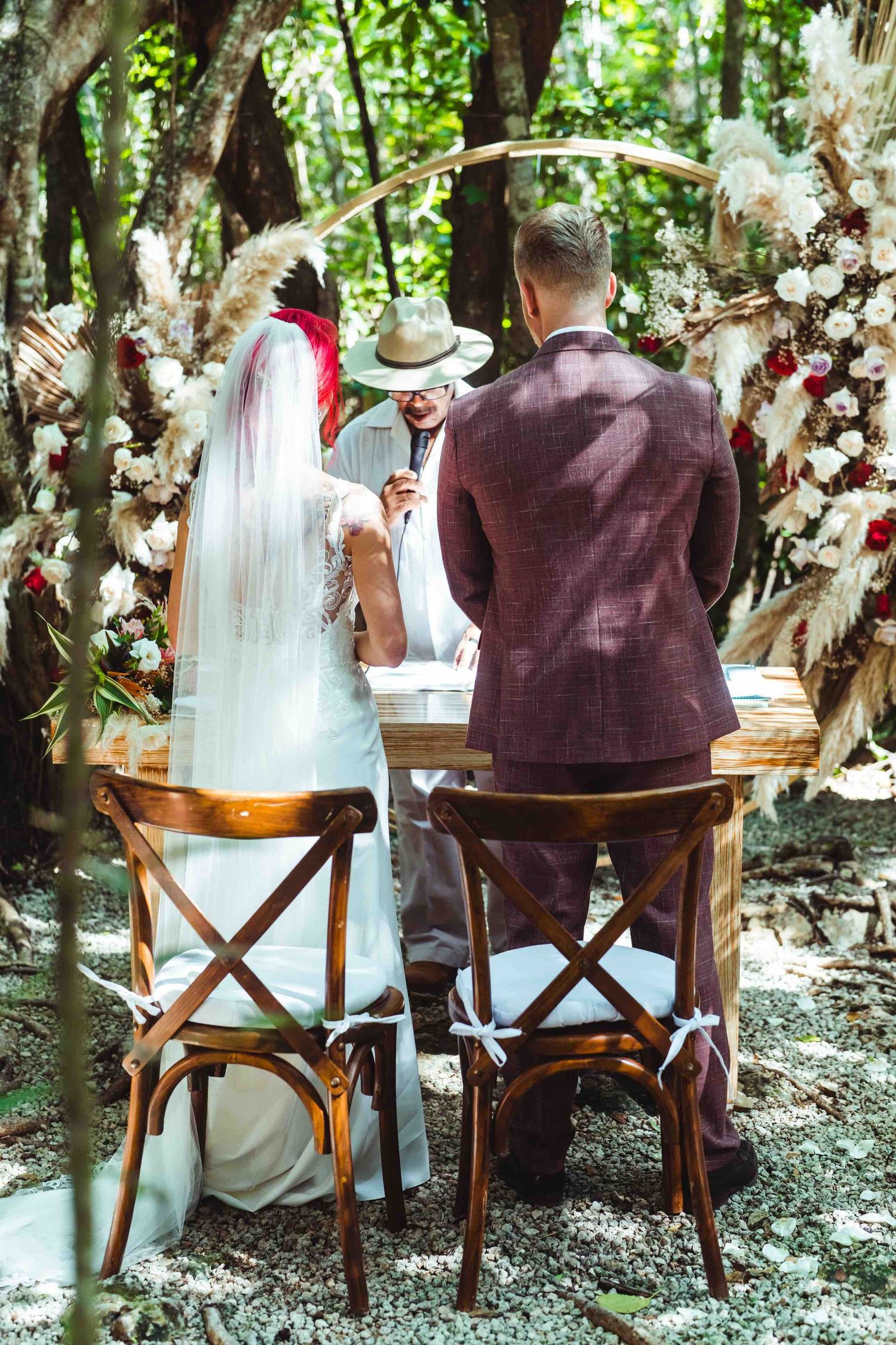 Bride and groom seated at wooden altar with floral arch during outdoor forest wedding ceremony.
