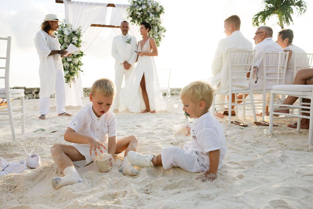 Two blonde children play in the sand during a beautiful beach wedding ceremony with white floral arch.