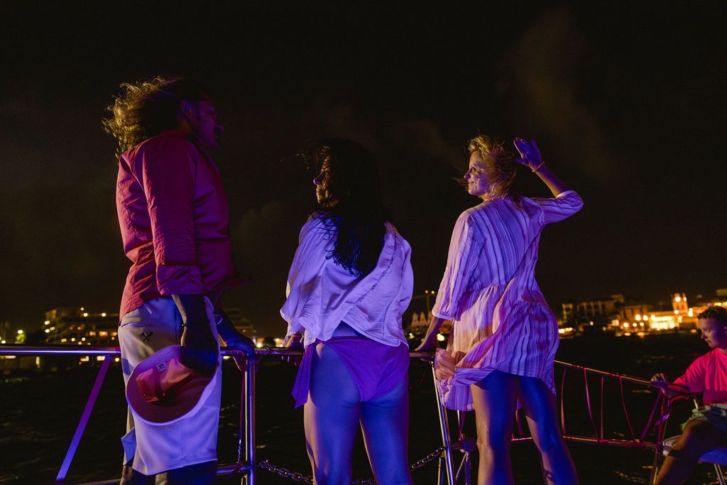Three women stand on a boat deck at night with city lights glowing in the background.