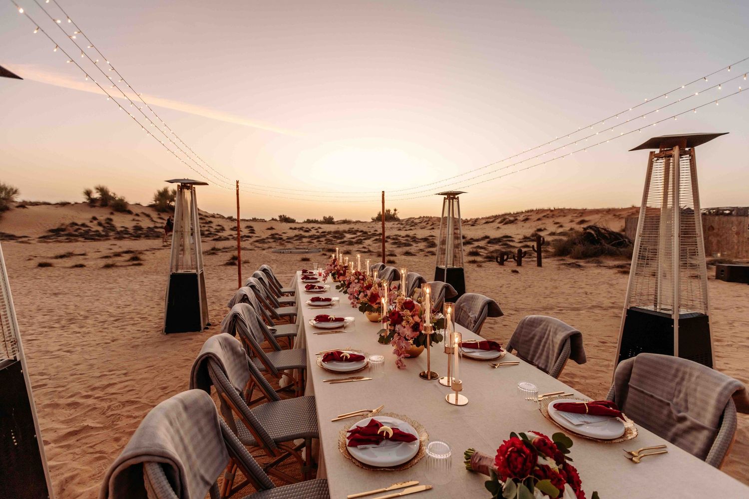Elegant long outdoor desert wedding reception table set with red floral centerpieces at golden sunset.