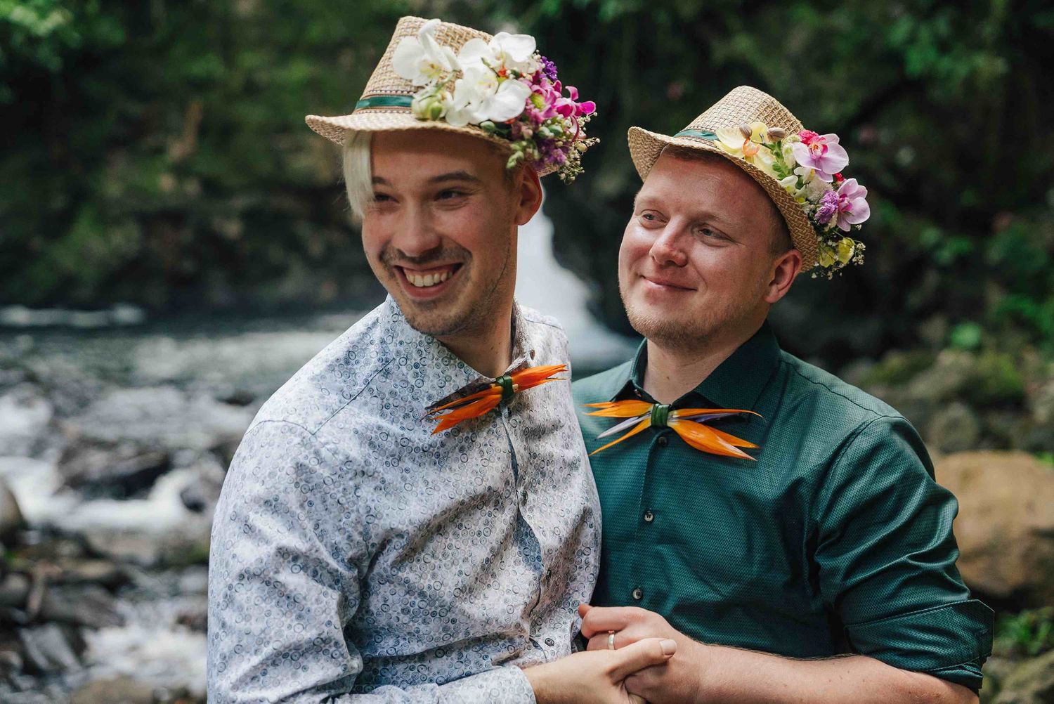 Two grooms wearing floral hats and colorful bow ties smile together near a waterfall in a lush forest setting.