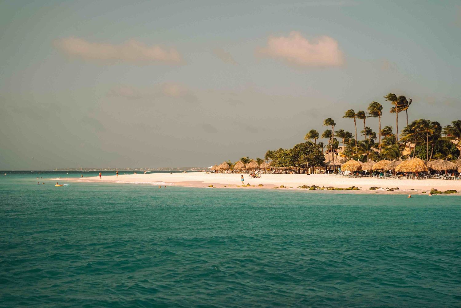 Turquoise Caribbean beach with white sand, palm trees, and thatched umbrellas under a partly cloudy sky.