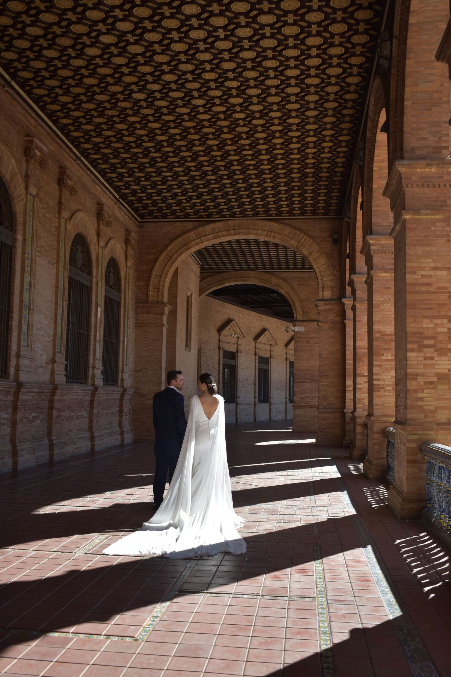 Bride and groom walk through a stunning ornate Spanish architectural corridor with dramatic light and shadows.