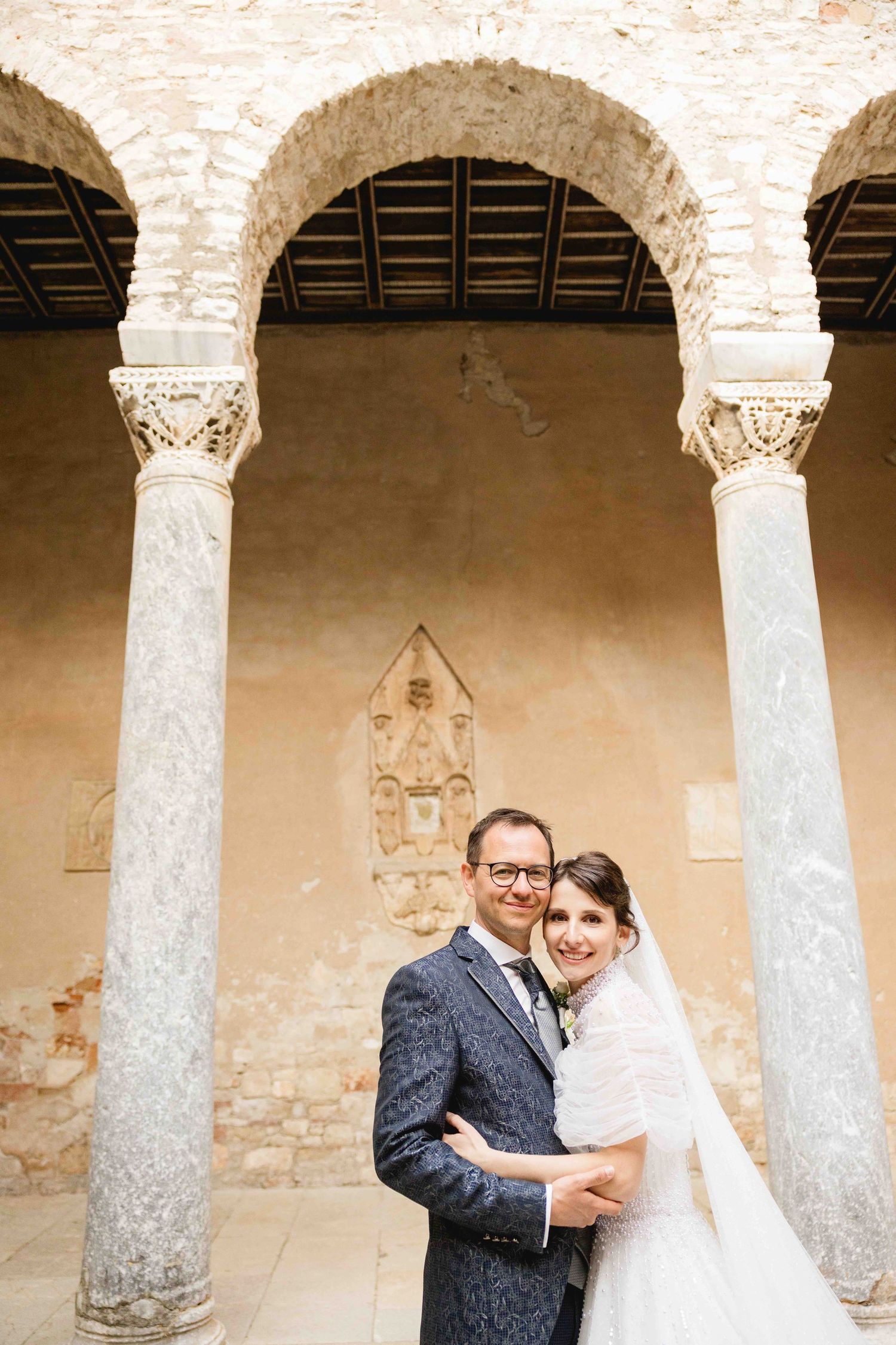 Wedding couple embracing under ornate stone arches with marble columns at a historic European venue.