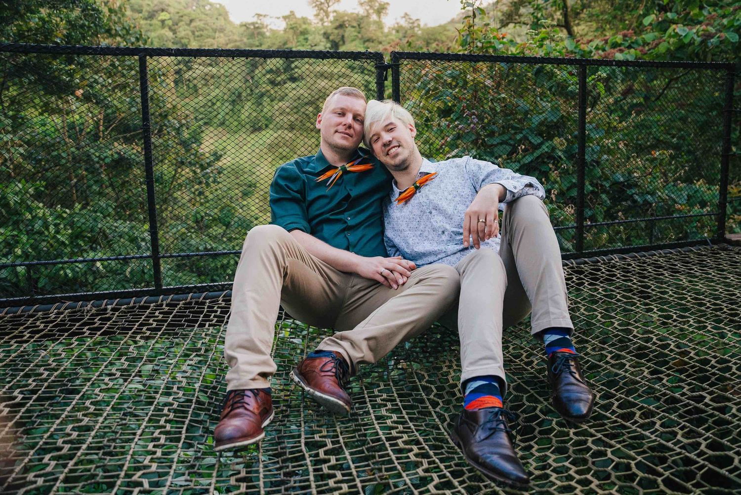 Two people sitting closely together on a net bridge surrounded by lush green trees, wearing colorful socks.