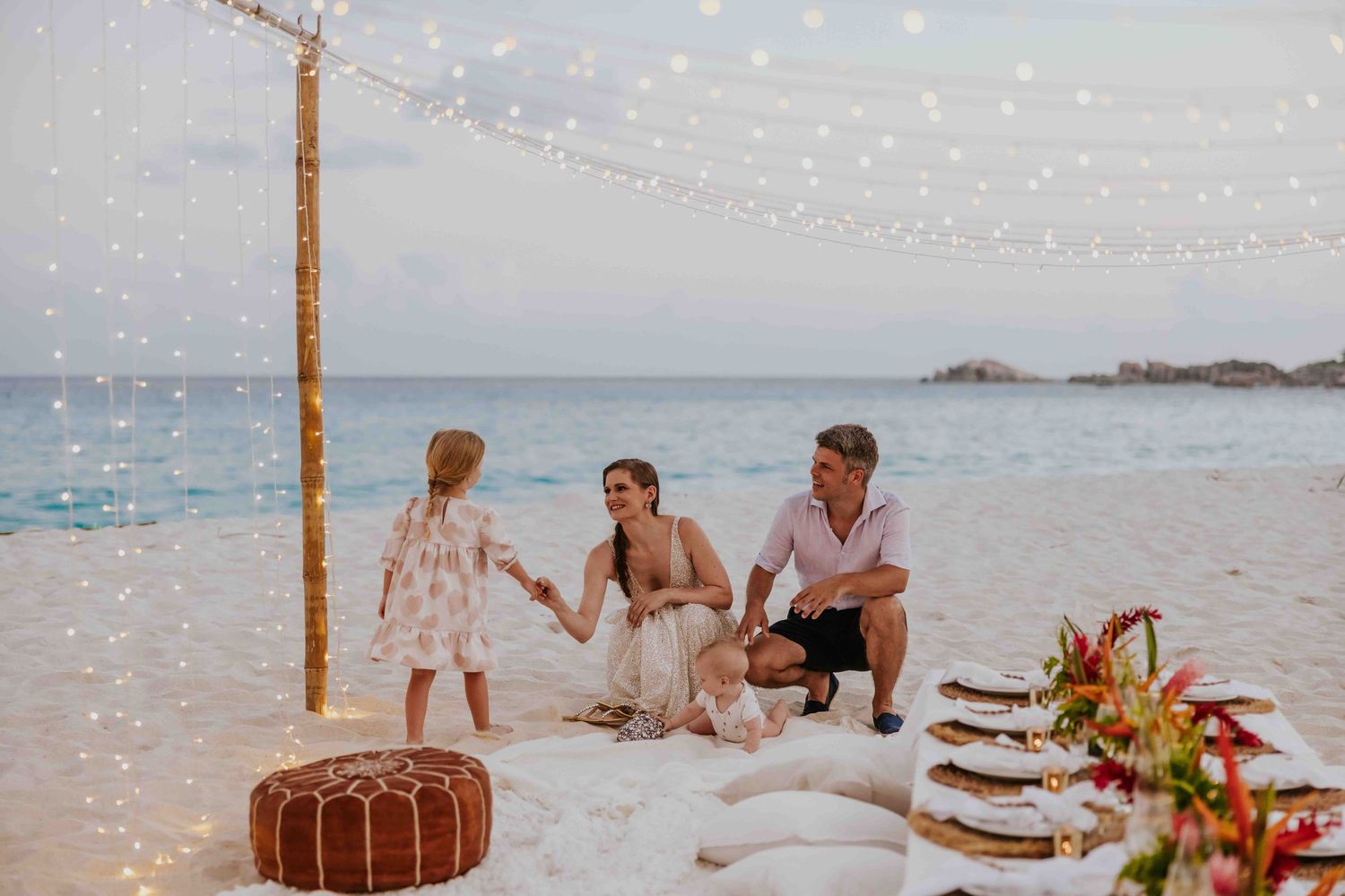 Family enjoying a romantic beach picnic setup with fairy lights at dusk by the ocean.